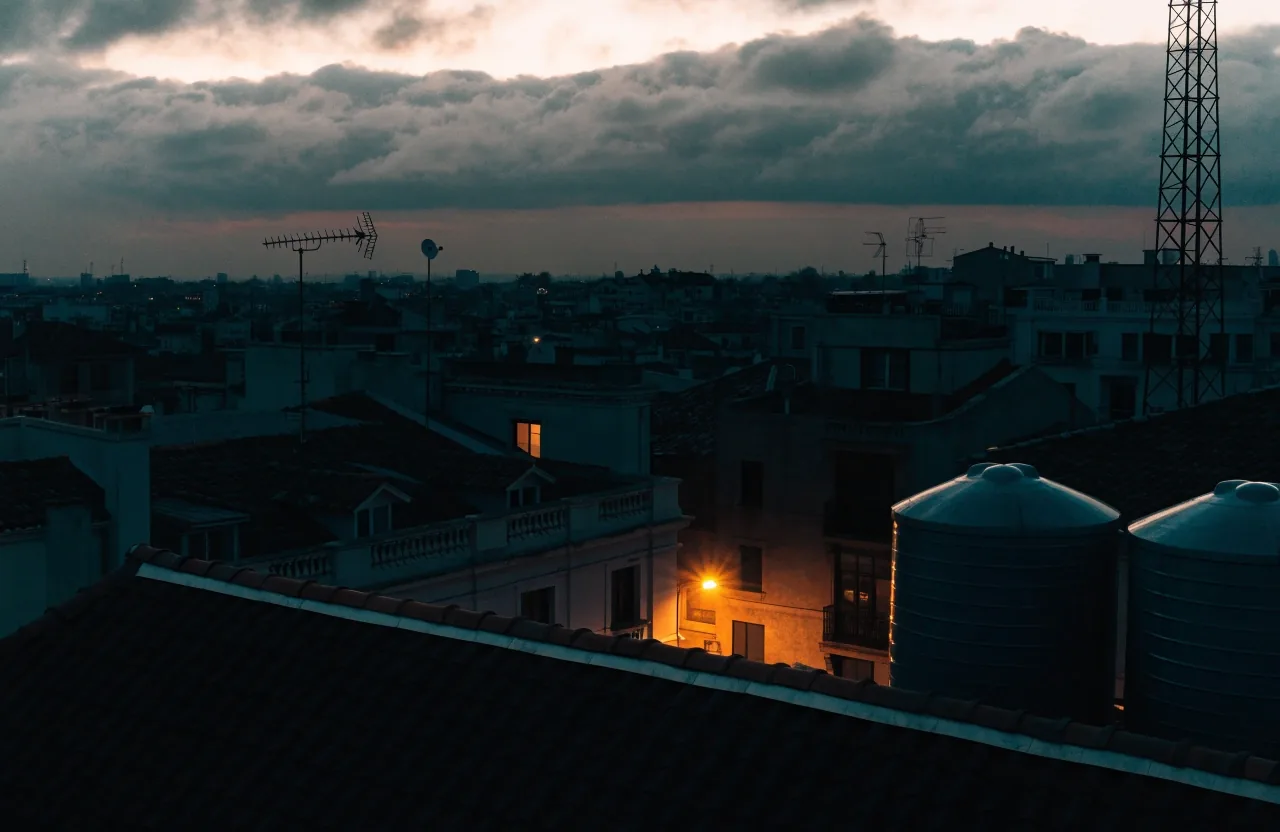 Tejados de una ciudad bajo nubes bajas al final del día, con una ventana iluminada y farolas encendidas